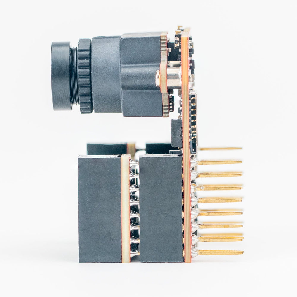 A compact electronic module with a black cylindrical lens-like component, dark gray rectangular blocks, gold-toned pins, and copper-colored circuitry, photographed on a clean white background.
