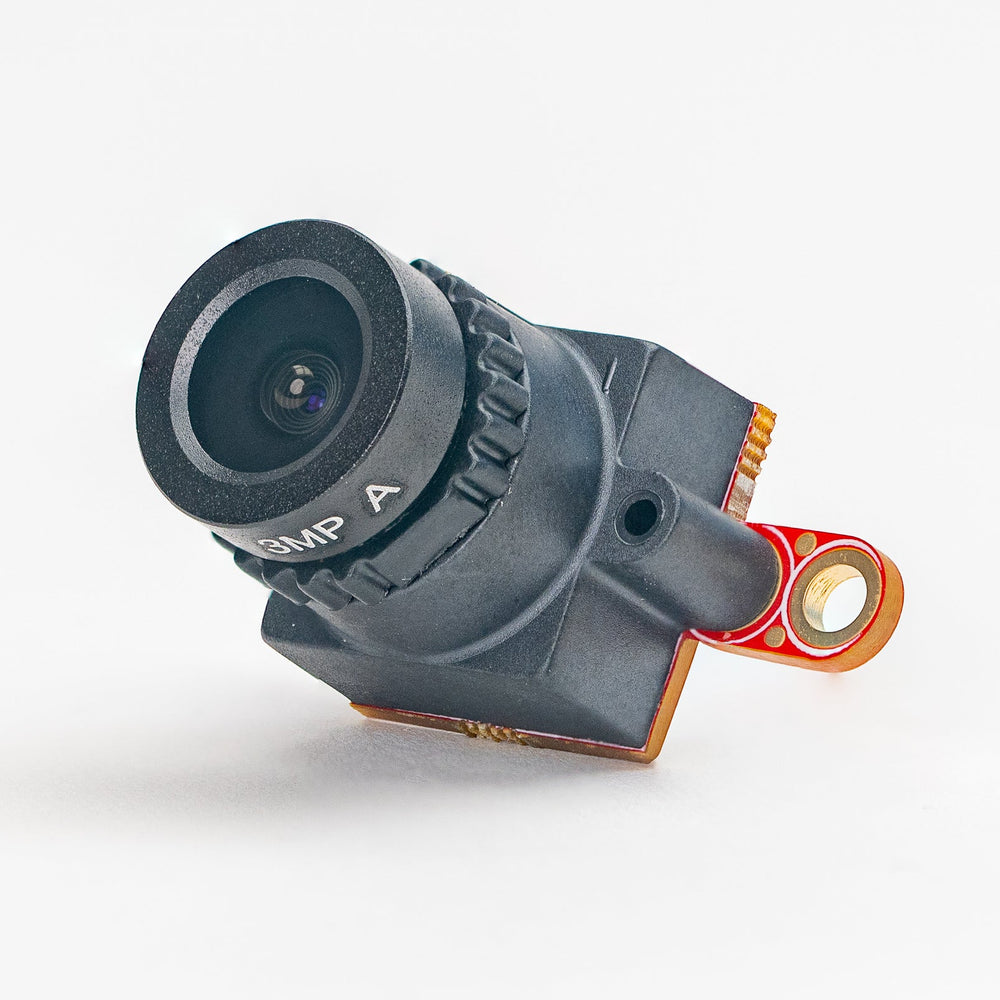 A miniature black FPV camera with a clear lens and orange accent wires, photographed on a seamless white background.